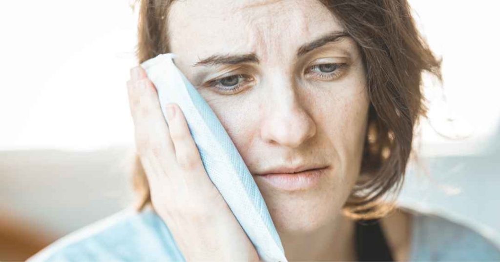 Woman wearing a light blue shirt holding an ice pack on her face to calm swelling down after a dental injury.