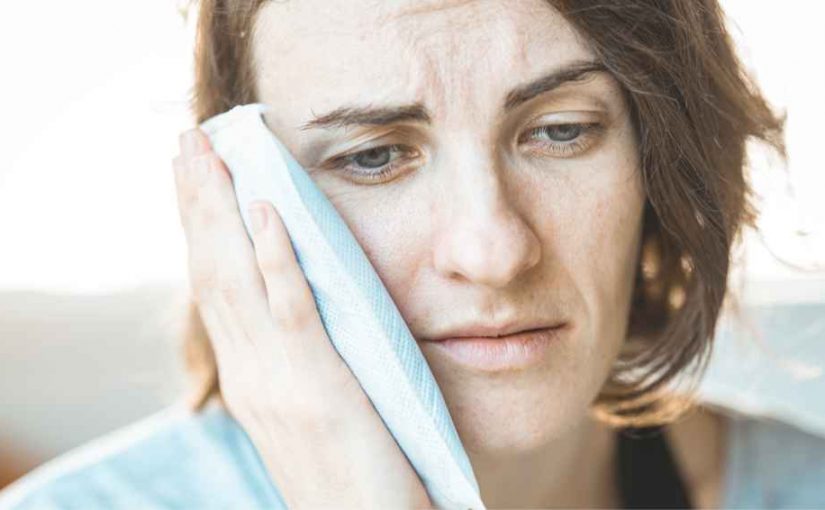 Woman wearing a light blue shirt holding an ice pack on her face to calm swelling down after a dental injury.